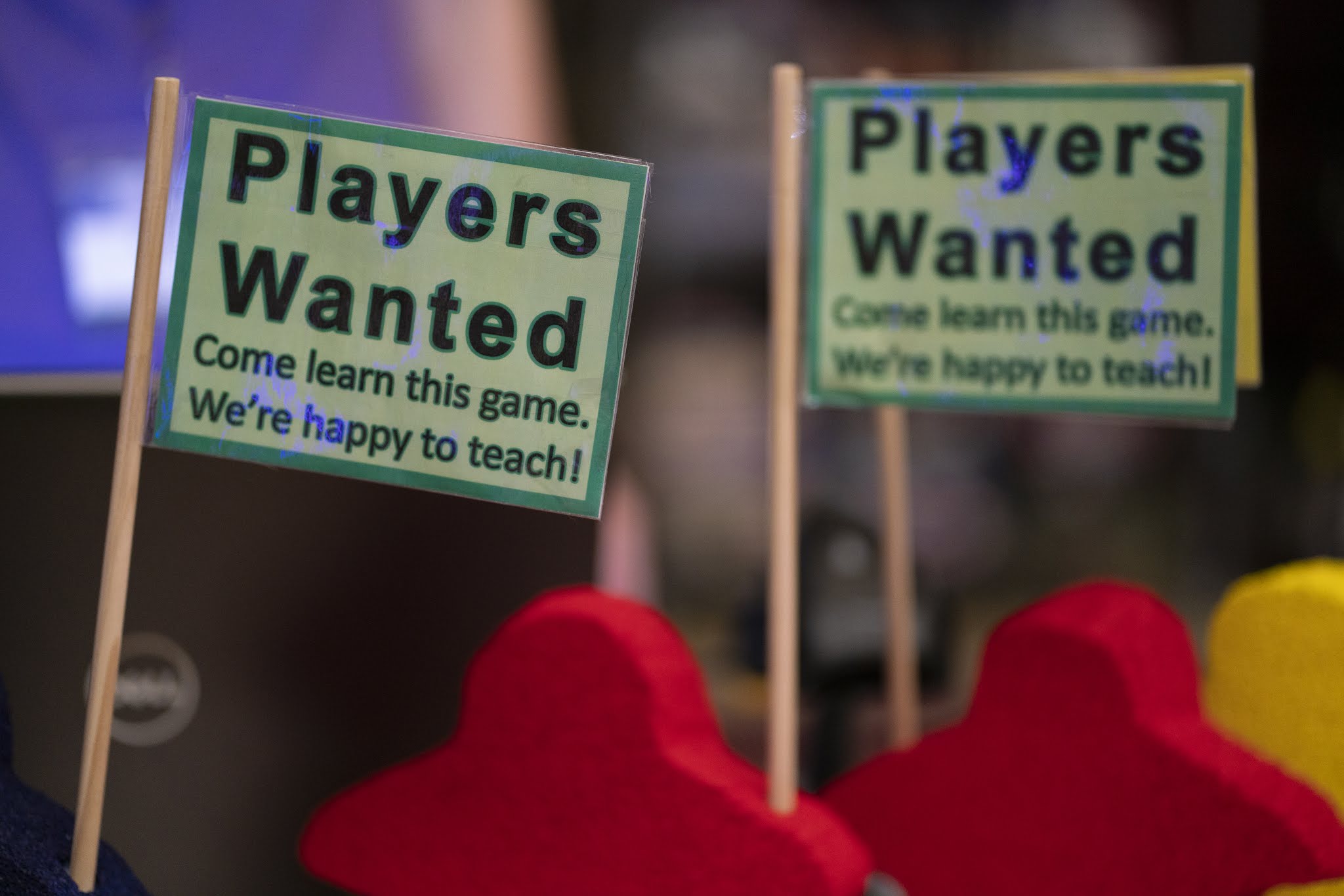 Meeple-shaped sign holders. Sign Reads "Players Wanted - Come Learn This Game. We're Happy to Teach!"